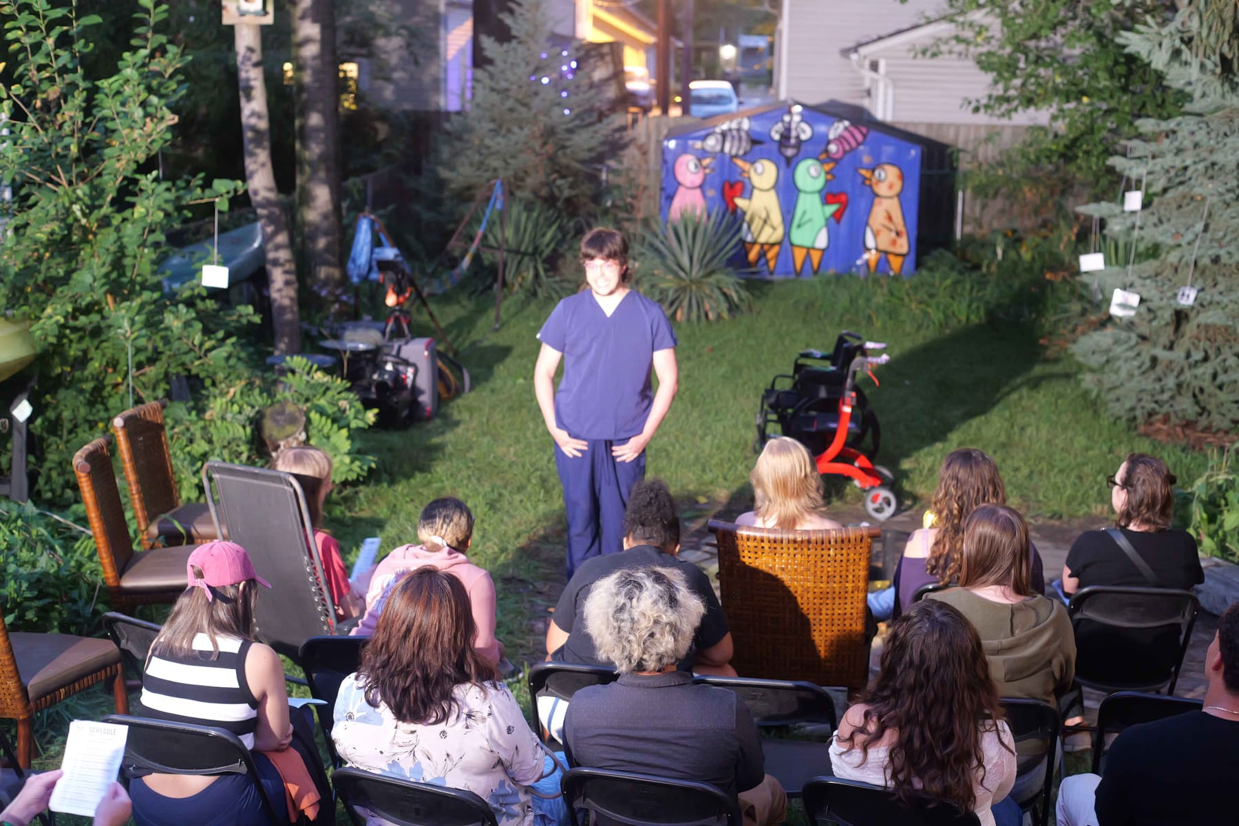 A performer in blue scrubs addresses an audience seated on mismatched chairs in a backyard, in front of a shed painted with cartoon birds