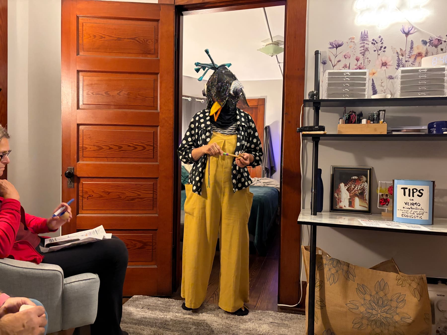 A masked bird character in a beaded headdress, polka-dot jacket, and yellow jumpsuit performs in a bedroom doorway for a seated audience