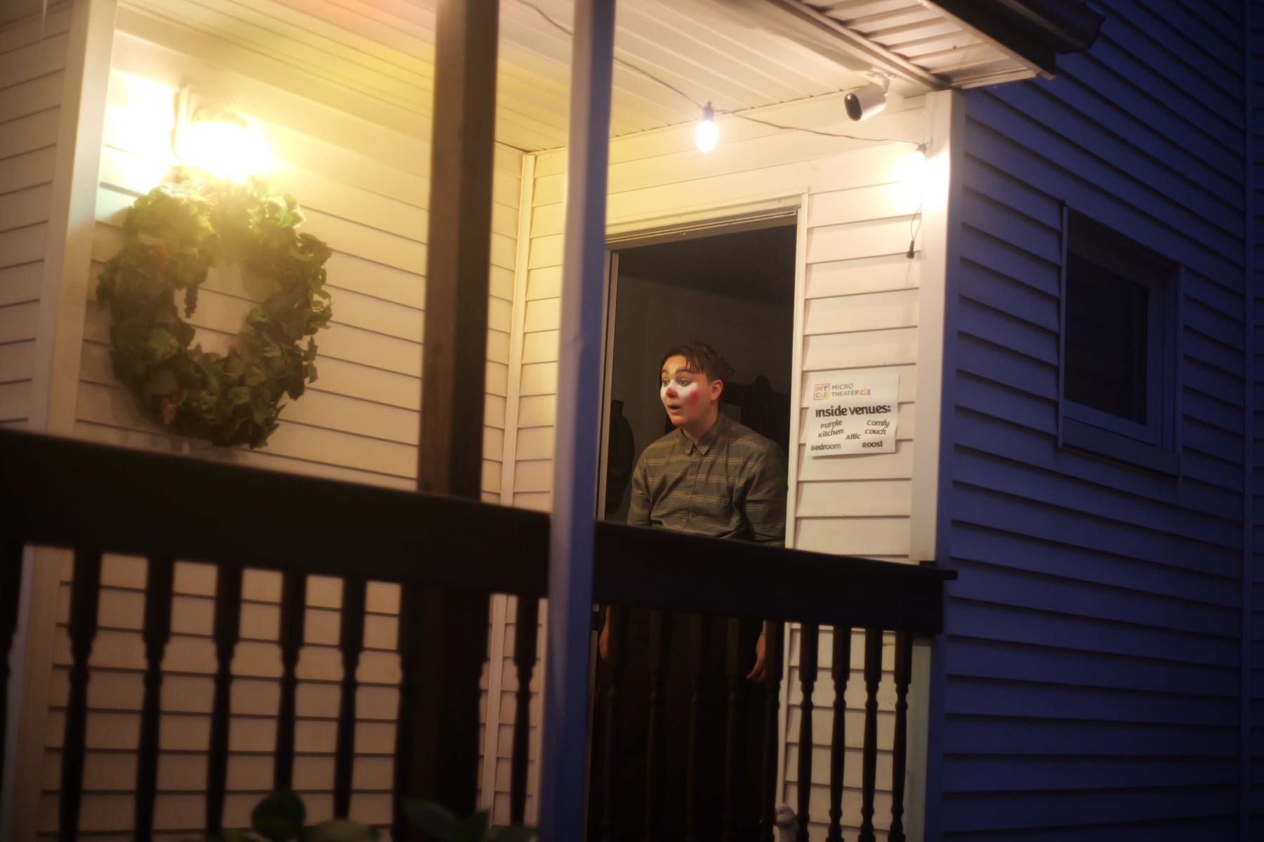 A performer in clown makeup leans out of a doorway on a porch at night, next to a sign listing the inside venues: Purple Kitchen, Comfy Couch, Attic, Bedroom, Roost