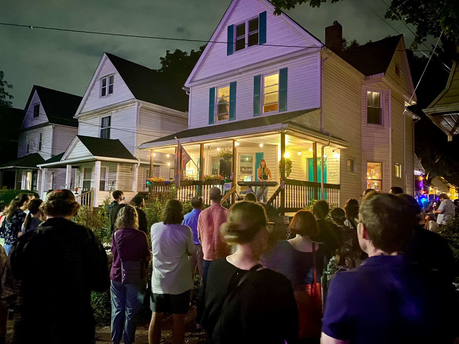 A singer at a keyboard performs on a string-lit front porch to a crowd standing in the yard and street at night